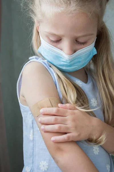 マスクの中の小さな女の子は 皮膚にプラスターが付いている腕を保持します ワクチン接種と予防接種の概念 — ストック写真