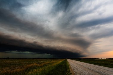 Issız bir yola doğru yaklaşan gök gürültülü bulutların dramatik bir görüntüsü. Düz tarlalar boyunca uzanan ağır gölgeler fırtınalı gerilimle dolu bir gökyüzü altında..