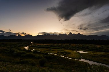Fırtına bulutları, gün batımı ışığı yer değiştirirken dağ manzarasında hareket eder. Gölge, doku ve renklerle dramatik bir gökyüzü yaratarak doğanın ve vahşi doğanın destansı ve dengesiz güzelliğini vurgular.