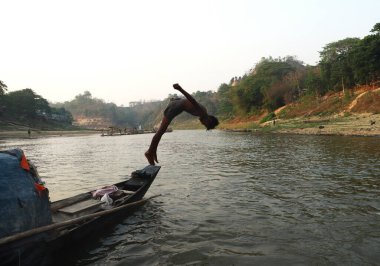 Bir çocuk arka planda ormanla birlikte tekneden nehre atlıyor. Çocuklar yüzüyor, zıplıyor ve günlük aktiviteler olarak oynuyor..