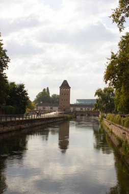 Sakin bir nehrin kenarında duran tarihi bir kule yansıması sakin sularda parıldıyor. İnsanlar yürüyüş yolunda yürürken tuğla setler ve yemyeşil alanlar nehir boyunca uzanır..
