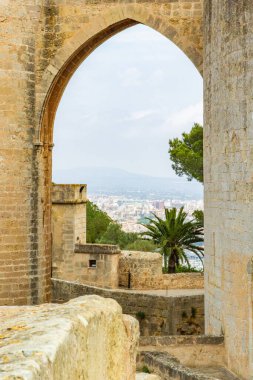Looking through and ancient stone arch there is a view of the city of Palma in the background, a palm tree in mid ground with blue sky in Mallorca Spain.
