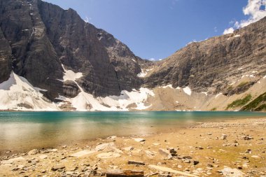 Buzul dağı, mavi gökyüzüyle sıcak güneşli bir günde kaya ve kumlu bir kıyı şeridiyle kristal berrak bir göle besleniyor.