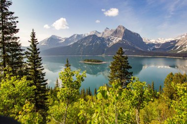 Arkaplanda, yeşil çalılar ve ağaçlarla çevrili yeşil bir gölün ortasında küçük bir adanın bulunduğu kar kaplı kayalık dağcıların manzarası.