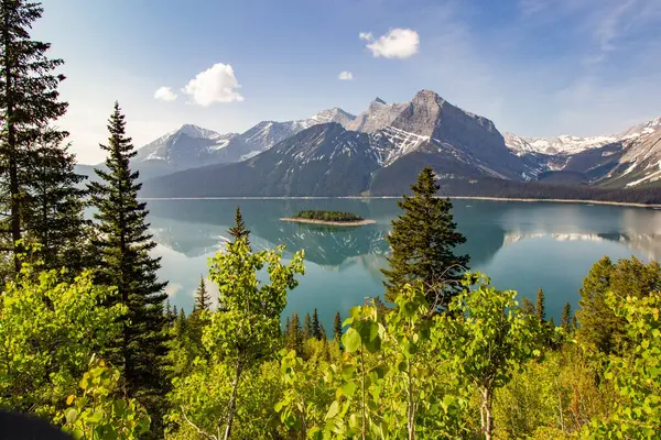Arkaplanda, yeşil çalılar ve ağaçlarla çevrili yeşil bir gölün ortasında küçük bir adanın bulunduğu kar kaplı kayalık dağcıların manzarası.