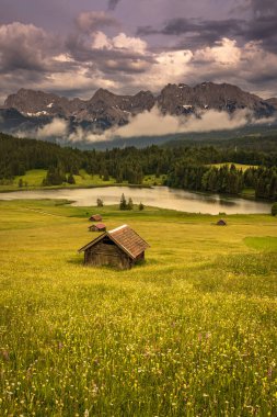 Ufukta karamsar bulutlar ve ağaçlar olan büyük bir dağı ve gölü gösteren küçük kulübelerin dikey görüntüsü.