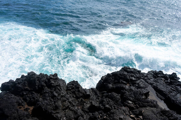 Idyllic scene of clear blue sea crashing against volcanic black rocks along the cliffs of Terceira Island, Azores.