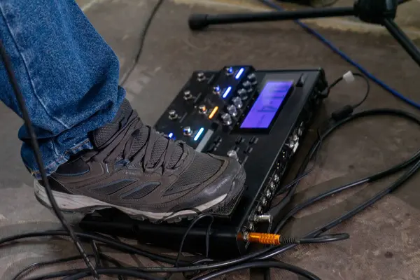 A foot steps on a guitar effects pedal, captured from the side. The shoe is a simple sneaker, and the pedal rests on a textured floor. The focus is on the action, with a sense of anticipation.