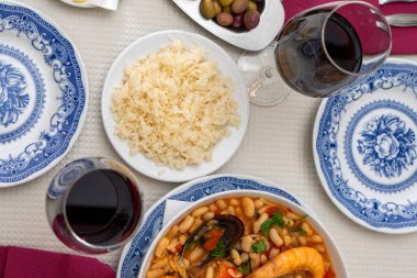 A dish of seafood feijoada with rice, garnished with shrimp and clams, sits on a restaurant table. Alongside are Alentejo cheese and a glass of red wine, evoking a warm Portuguese dining experience. clipart