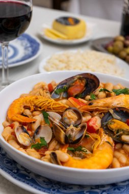 A dish of seafood feijoada with rice, garnished with shrimp and clams, sits on a restaurant table. Alongside are Alentejo cheese and a glass of red wine, evoking a warm Portuguese dining experience. clipart