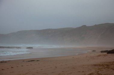 Portekiz, Aljezur 'daki Monte Clrigo plajları sabah sisi ile kaplanarak manzarayı yumuşatıyor. Yumuşak dalgalar kıyıya vurur, dingin ve mistik bir sahil manzarası yaratır..