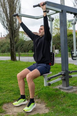 Gri saçlı bir adam, şehir parkındaki bir spor salonunda göğüs pres makinesinde çalışıyor. Kararlılıkla egzersiz yapar, temiz havanın tadını çıkarır ve aktif kalır..