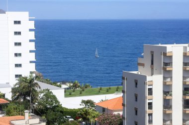 Madeira Adası Funchal 'daki iki bina arasında zarif bir şekilde süzülen küçük bir yelkenli, berrak bir gökyüzü ve parıldayan sular tarafından çerçevelenmiş, sakin bir denizcilik anını yakalıyor..