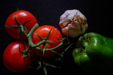 A vibrant still life of three ripe tomatoes, a green bell pepper, a garlic bulb, and an onion on a black background, highlighting their fresh textures and colors. clipart