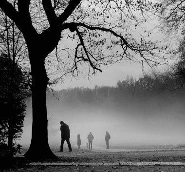 Sisli bir parkta ağaçların arasında yürüyen insanların atmosferik siyah beyaz bir fotoğrafı doğal manzarayı ve eşsiz atmosferik fenomeni gözler önüne seriyor.