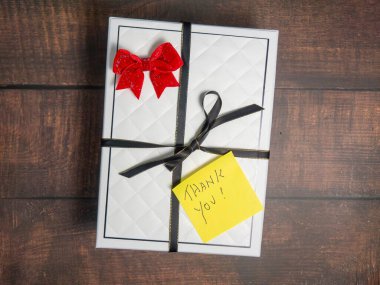 Sarı teşekkür notu. Üzerinde kırmızı fiyonk ve siyah kurdele olan beyaz bir hediye kutusu var.