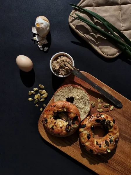 stock image A snack of bagels with pate and eggs. Bagel on cutting board. Pate with a knife in a small bowl and boiled eggs are nearby. A sprouting onion on a pot holder. 