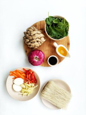 Ingredients for preparing the Korean dish Chapche. Vegetarian cooking method. Sweet pepper, hot pepper, white onion and rice noodles in plates. Mushrooms, spinach, onion, soy sauce and sesame oil on a cutting board. White background. clipart