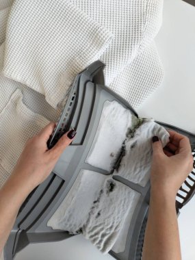 In the background is a dried, white robe, and next to it is a clothes dryer filter. The filter is filled with dust, hair and animal fur. Female hands taking the lint out from the filter of a drying machine.  clipart