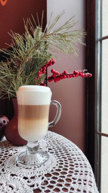 Layered latte in a clear glass mug with creamy foam, set on a crocheted tablecloth. Festive decor with pine branches and red berries by a window creates a cozy, holiday-inspired atmosphere. clipart