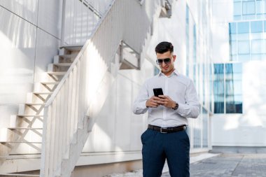 Beyaz gömlek ve mavi pantolon giyen bir adam cep telefonu ekranına odaklanmış durumda. Açık havada duruyor gibi görünüyor, muhtemelen şehir ortamında..