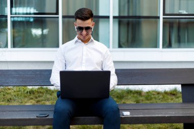 Dışarıdaki ahşap bir bankta oturan bir adam dizüstü bilgisayarına oturmuş daktilo yazıyor. Arka planda ağaçlar ve yeşillikle işine odaklanmış ve meşgul görünüyor..