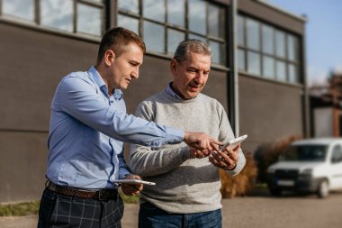 Güneşli bir günde iki adam bir binanın önünde dikiliyorlar. Mavi gömlekli ve ekose pantolonlu genç bir adam yaşlı adamın elindeki tableti işaret ediyor. Yaşlı adam gri bir kazak ve kot pantolon giyiyor ve tablete bakıyor..