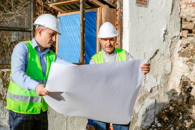 Beyaz kasketli ve yeşil yelekli iki inşaat işçisi, kısmen yıkılmış bir binanın önünde durup planları inceliyor. Adamlar planları gözden geçirirken konuşuyorlar..