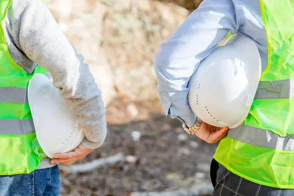 stock image Two construction workers, wearing green safety vests and holding white hard hats, stand side-by-side in front of a rocky hillside. The workers are positioned in a way that their torsos are facing away from the camera, and their arms are bent, holding