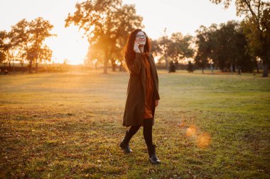 Uzun paltolu bir kadın parkta yürüyor, güneş batarken telefonla konuşuyor..