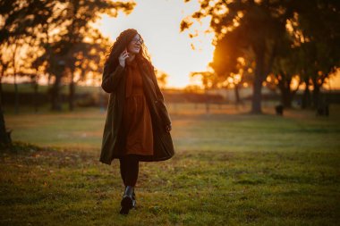 Uzun paltolu bir kadın gün batımında çimenli bir parkta yürüyor. Altın ışık, o ufka doğru yürürken sahneyi aydınlatıyor..