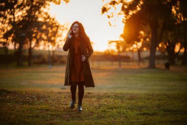 Paltolu ve gözlüklü bir kadın cep telefonuyla konuşurken parkta yürüyor. Güneş batıyor, sahne üzerine sıcak bir parıltı saçıyor..