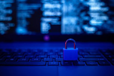 Mavi bir asma kilit, mavi bir parıltıyla aydınlatılmış bir dizüstü bilgisayarın klavyesinde duruyor. Ekran, veri güvenliğini vurgulayarak bulanık kod görüntüler.