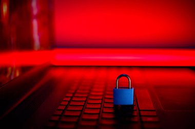 Mavi bir asma kilit dizüstü bilgisayarın klavyesinde duruyor. Arka planda kırmızı ışık yanıyor..