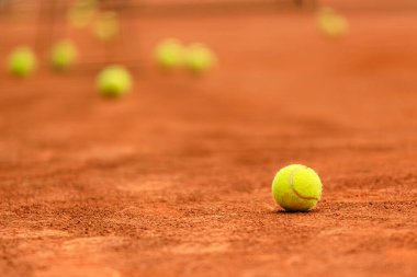Bir tenis topu, yüzeye dağılmış diğer toplarla çevrili bir tenis kortunun kırmızı kiline bağlıdır. Güneş ışıl ışıl parlıyor, kortta sıcak bir parıltı yayıyor..