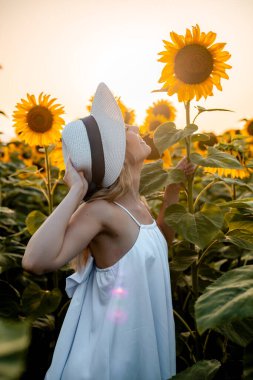 Beyaz elbiseli ve şapkalı bir kadın gün batımında tarlada büyük bir ayçiçeğine bakarken gülümsüyor..