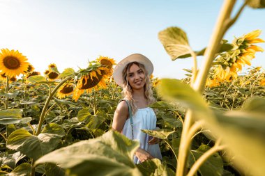 Beyaz bir elbise ve hasır şapka giyen genç bir kadın ayçiçeği tarlasında dururken gülümsüyor..
