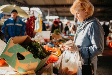 Bir kadın market tezgahında elinde plastik torba ve diğer şeyleri tutarken bir kutu yapraklı yeşili inceliyor..