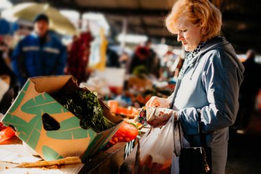 Mavi ceketli bir kadın markette plastik poşet tutarken ürünün fiyatına bakar..