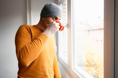 Gri bereli ve turuncu kazaklı bir adam pencerenin yanında duruyor, elinde kırmızı bir kupayla dışarı bakıyor..