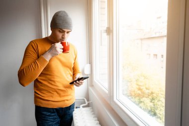 Örgü şapka takan bir adam kahve içer ve pencerenin yanında dikilirken telefonunu kontrol eder..