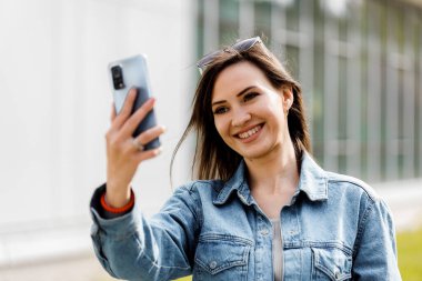 Uzun kahverengi saçlı ve güneş gözlüklü bir kadın akıllı telefonuyla gülümserken selfie çekiyor..