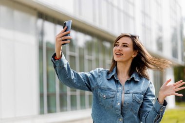 Kot ceketli genç bir kadın akıllı telefonuyla selfie çekerken ışıl ışıl gülümsüyor. Şehir zeminiyle birlikte açık havada..