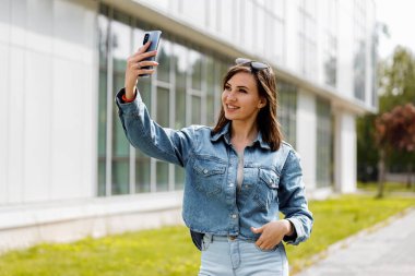 Bir kadın güneşli bir günde modern bir binanın önünde yürürken akıllı telefonuyla selfie çeker..