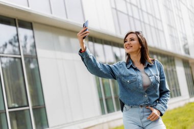 Bir kadın güneşli bir günde modern bir binanın önünde yürürken akıllı telefonuyla selfie çeker..