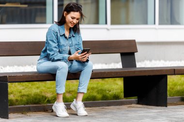 Dışarıda bir bankta oturan genç bir kadın telefonuna bakarken gülümsüyor..