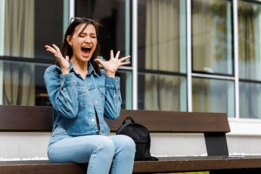 Kot ceketli genç bir kadın bankta oturuyor ve elleri havada şaşkınlıkla bağırıyor..