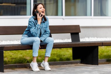 Binanın dışındaki bankta oturan bir kadın cep telefonuyla konuşuyor ve gülüyor..