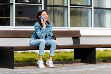 Kot ceket ve kot pantolon giyen genç bir kadın modern bir binanın önündeki bankta oturmuş akıllı bir telefonla konuşuyor..