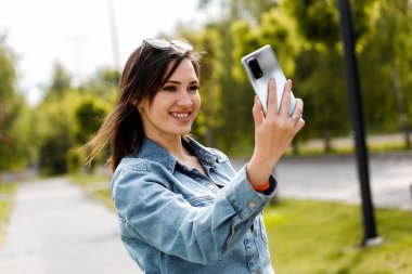 Kot gömlekli bir kadın güneşli bir günde parkta dikilirken akıllı telefonuyla selfie çekiyor..